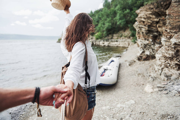 Portrait of happy young woman in hat holding man by hand and going to the boat on the lake in the mountains. - Photo, image
