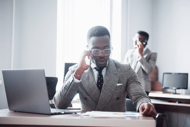Business call. Young african-american businessman in white has mobile phone talk in modern white office interior. - 写真・画像
