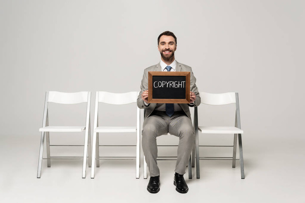 šťastný podnikatel drží tabuli se slovem copyright zatímco sedí na židli na šedém pozadí - Fotografie, Obrázek