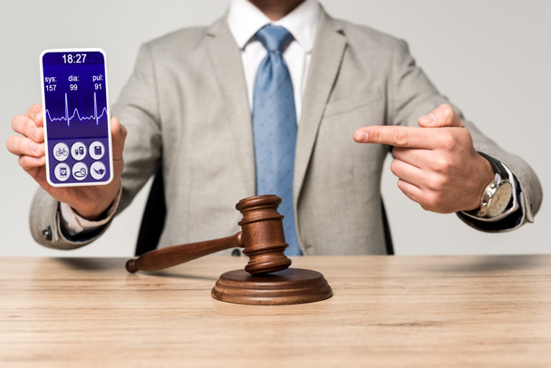 oříznutý pohled na právníka ukazujícího prstem na smartphone s aplikací tepové frekvence a soudcovskou kladívko na stole - Fotografie, Obrázek