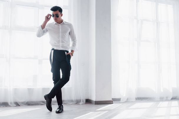 Sits perfect. Full length of beautiful young man adjusting his sunglasses in the white room. - Foto, Imagem