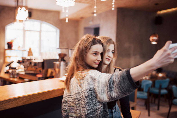 Two friends drinking coffee in a cafe, taking selfies with a smart phone and having fun making funny faces. Focus on the girl on the left. - Foto, imagen