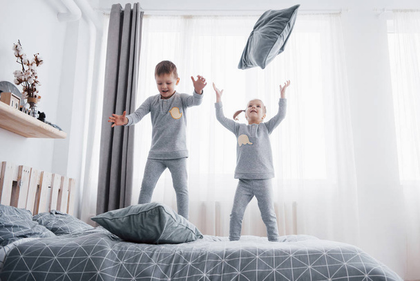 Happy kids playing in white bedroom. Little boy and girl, brother and sister play on the bed wearing pajamas. Nightwear and bedding for baby and toddler. Family at home. - Zdjęcie, obraz