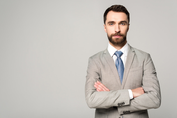 confident businessman standing with crossed arms and looking at camera isolated on grey - Foto, afbeelding