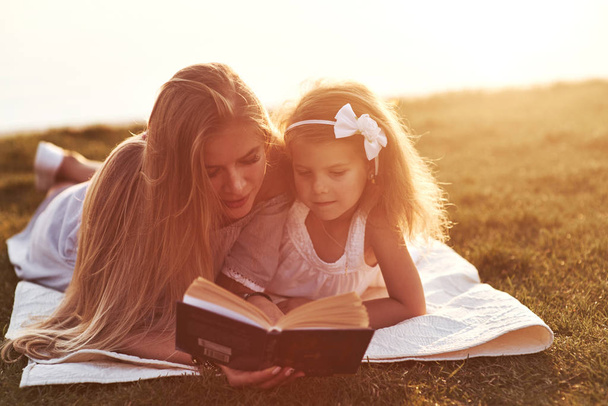 That piece of art got attention. Mom and daughter reading a book in a very sunny day laying on the grass with lake at background. - Photo, Image