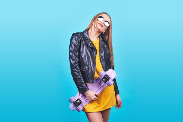 Attractive tanned girl with smile in face wears black leather jacket posing with pleasure. Indoor photo of lovely woman in sunglasses standing with skateboard on blue background. - 写真・画像