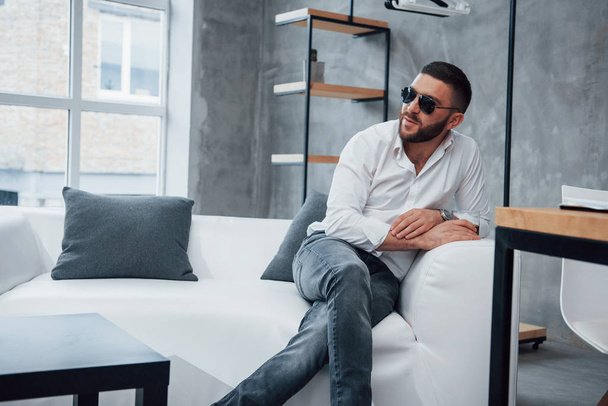 Young short-haired man in sunglasses sitting on couch in the office. - Фото, изображение