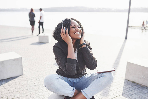 Portrait of a beautiful young pretty African American girl sitting on the beach or lake and listening to music in her headphones. - Fotografie, Obrázek