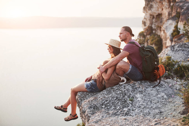 Hugging couple with backpack sitting on top of rock mountain enjoying view coast a river or lake. Traveling mountains and coast, freedom and active lifestyle concept. - Photo, image