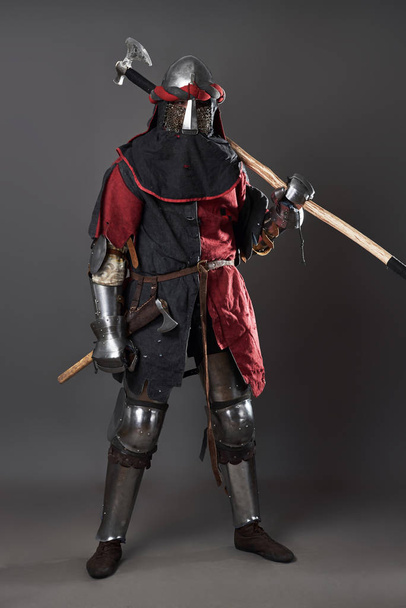 Medieval knight on grey background. Portrait of brutal dirty face warrior with chain mail armour red and black clothes and battle axe. - Valokuva, kuva