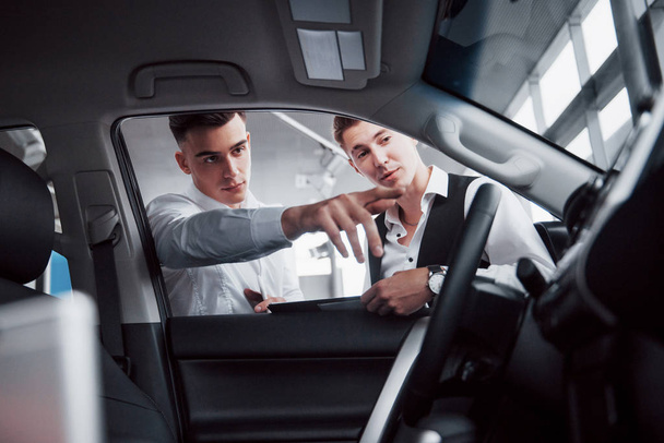 Twee mannen staan in de showroom tegen auto 's. Close-up van een sales manager in een pak dat een auto verkoopt aan een klant. - Foto, afbeelding
