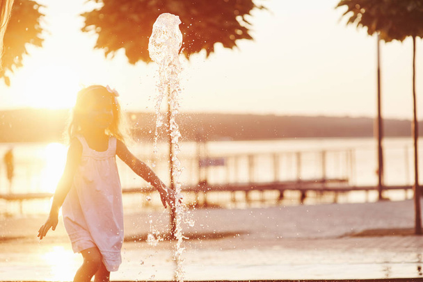 Young girl play in the fountain at the summer heat and lake and woods background - Foto, Imagem