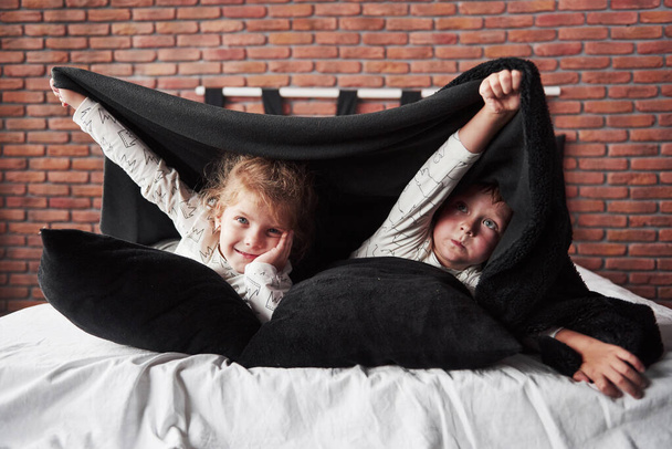 Küçük çocuklar, kız ve erkek battaniyeyle yatak örtüsünün üzerinde yastıklarla yatıp oynuyorlar.. - Fotoğraf, Görsel