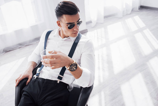 Handsome guy. Young stylish businessman sitting on a black chair with glass of whiskey. - 写真・画像