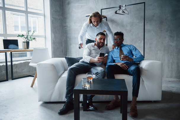 Man with short hair showing interesting information on smartphone. Group of multiracial office workers in formal clothes talking about tasks and plans. - Fotoğraf, Görsel