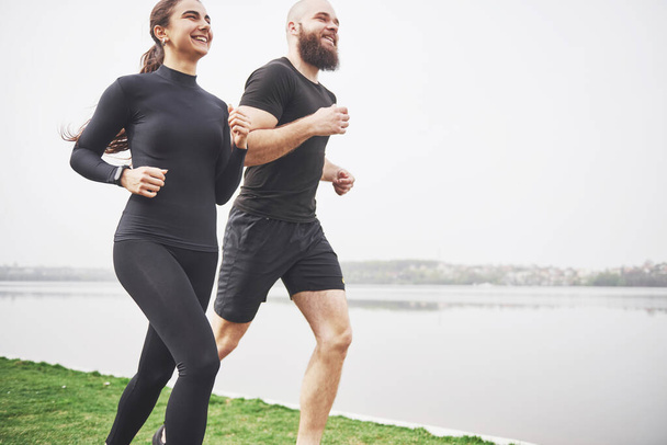 Couple jogging and running outdoors in park near the water. Young bearded man and woman exercising together in morning. - Фото, изображение