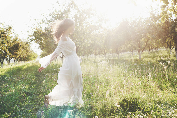 Beautiful young woman wearing elegant white dress and enjoying beautiful sunny afternoon in a summer garden. - Φωτογραφία, εικόνα