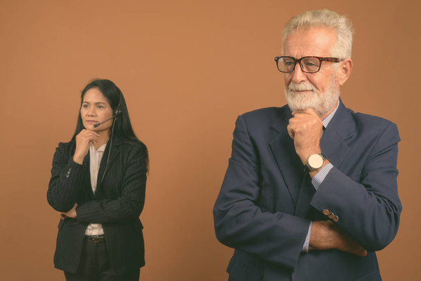 Студийный снимок старшего бизнесмена и зрелой азиатской бизнесвумен вместе на коричневом фоне
 - Фото, изображение