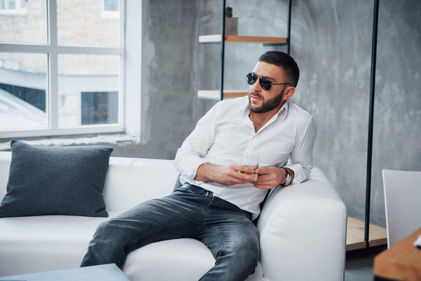 Thoughts about future. Young short-haired man in sunglasses sitting on couch in the office. - Foto, Bild