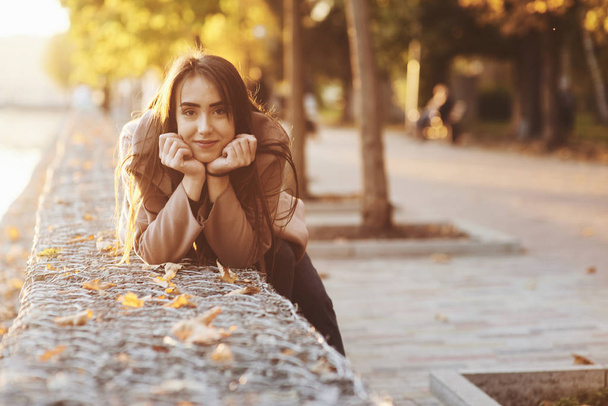 Young, pretty brunette girl leaned on the wall with her two hands under the chin, wearing casual coat with an autumn sunny park on the blurry background with a copy space. - Фото, изображение