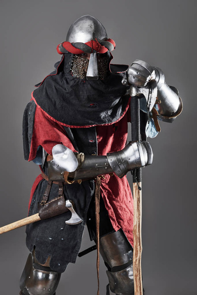 Medieval knight on grey background. Portrait of brutal dirty face warrior with chain mail armour red and black clothes and battle axe. - Zdjęcie, obraz