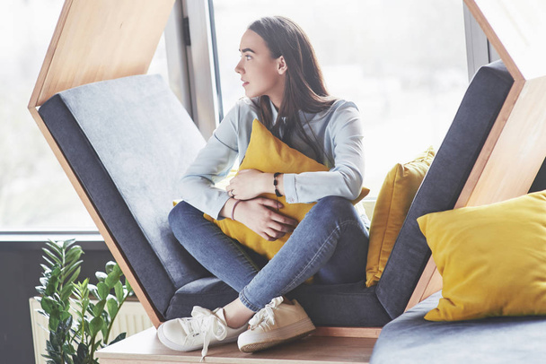 Beautiful young girl spends her time in the recreation area. She sits and has fun in the sofa chair in the form of a hexagon with pillows near the window. - Foto, Bild