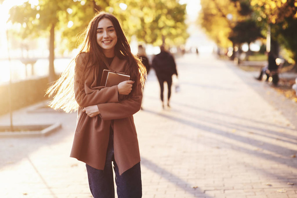Front view of young, smiling, pretty brunette girl standing and holding small stack of books in her hands, wearing casual coat at autumn sunny park alley on blurry background with a copy space. - Valokuva, kuva