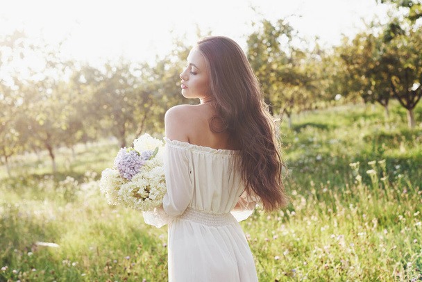 A beautiful young girl in a white light dress and a bouquet of summer flowers lays a fine day in the garden. - Valokuva, kuva