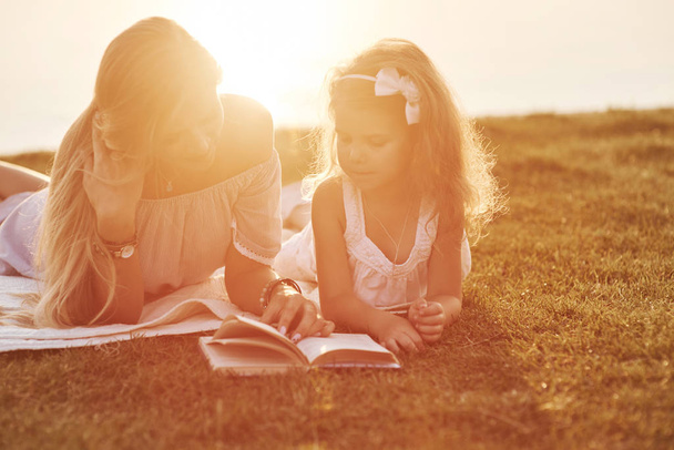 Mom and daughter reading a book in a very sunny day laying on the grass with lake at background. - 写真・画像