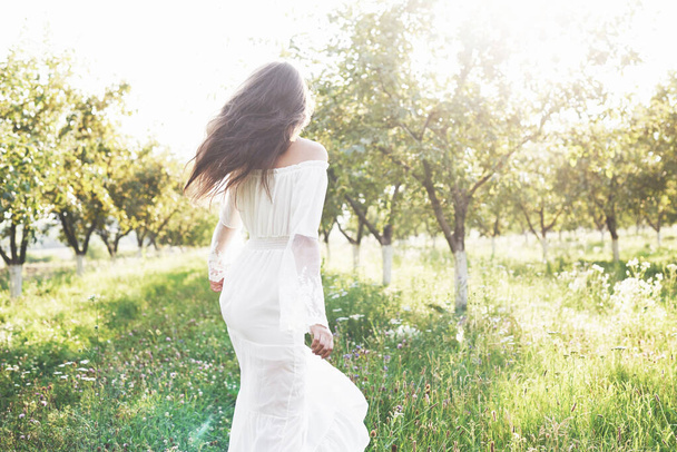 A young girl in a white long dress is walking in the garden. Beautiful sunset through the leaves of trees. - Zdjęcie, obraz