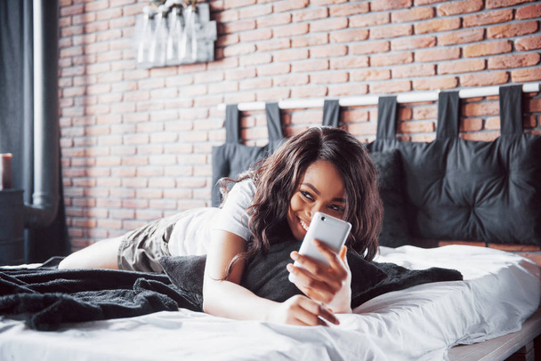 Portrait of beautiful woman waking up in her bed and looks into the phone. Check social networks, send sms. The girl is wearing a T-shirt. - Zdjęcie, obraz
