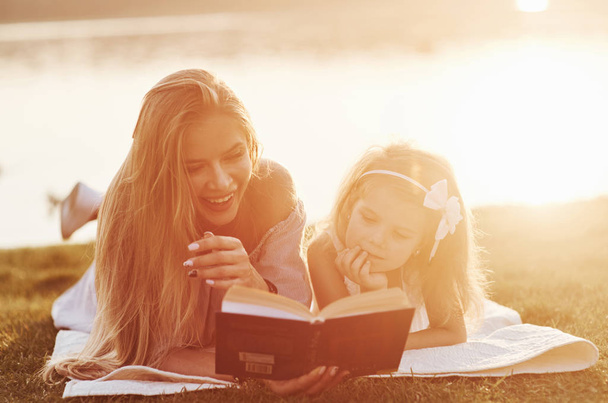 Stumbled upon a joke. Mom and daughter reading a book in a very sunny day laying on the grass with lake at background. - Fotografie, Obrázek