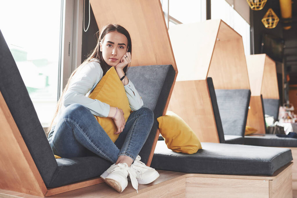 Beautiful young girl spends her time in the recreation area. She sits and has fun in the sofa chair in the form of a hexagon with pillows near the window. - Foto, afbeelding