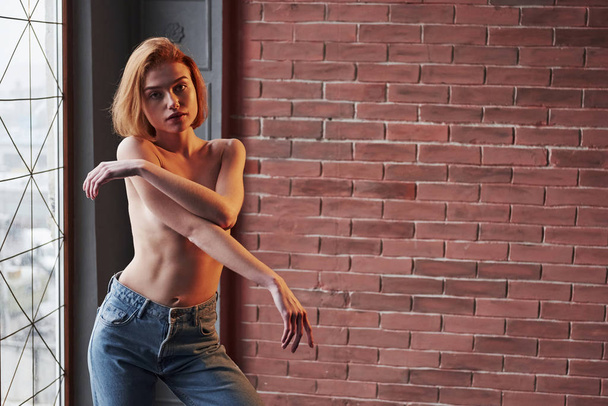 Erotical portrait. Hot young blonde with bare chest and jeans stands against the window and brick wall. - Φωτογραφία, εικόνα