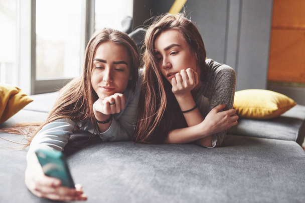 Two cute smiling twins sisters holding smartphone and making selfie. Girls lie on the couch posing and joy. - Фото, изображение