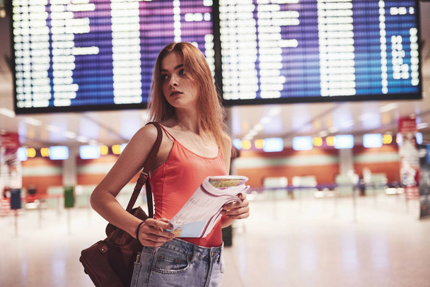 Beautiful young tourist girl with backpack in international airport, near flight information board. - Foto, Imagen