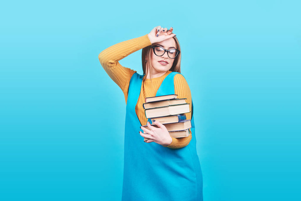 Confused cute girl in glasses holding in hands a pile of books isolated on colorful blue background. - Photo, Image