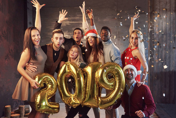 New 2019 Year is coming! Group of cheerful young multiethnic people in Santa hats carrying gold colored numbers and throwing confetti on the party. - Foto, afbeelding
