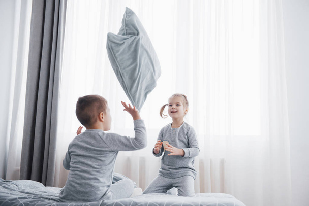 Happy kids playing in white bedroom. Little boy and girl, brother and sister play on the bed wearing pajamas. Nursery interior for children. Nightwear and bedding for baby and toddler. Family at home - Valokuva, kuva