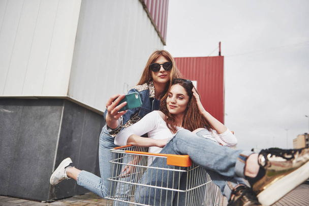 Two young happy women having fun shopping trolley race outdoors and make sephi - Valokuva, kuva