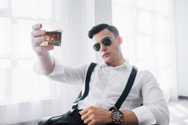 Good lighting. Young stylish businessman in sunglasses sitting on a black chair with glass of whiskey. - Valokuva, kuva
