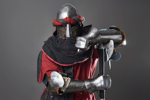 Medieval knight on grey background. Portrait of brutal dirty face warrior with chain mail armour red and black clothes and battle axe. - Fotografie, Obrázek