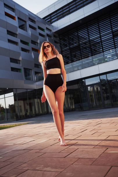 Sexy young woman in sunglasses and black bathing suit is standing on the city buildings background during sunset. - Zdjęcie, obraz
