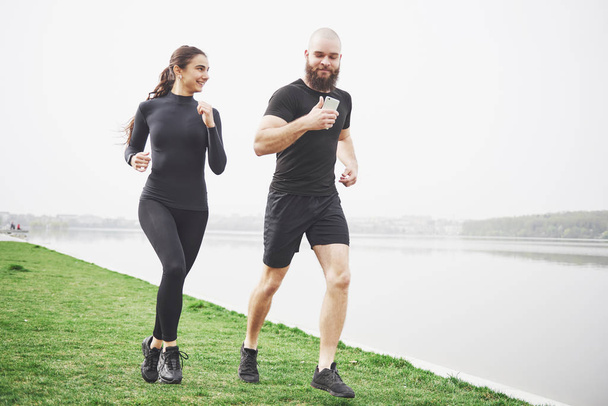 Couple jogging and running outdoors in park near the water. Young bearded man and woman exercising together in morning. - Fotó, kép