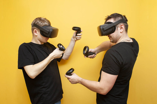 two guys in VR glasses on a yellow background play a shooter, friends gamers rivals in virtual reality glasses fight - Valokuva, kuva