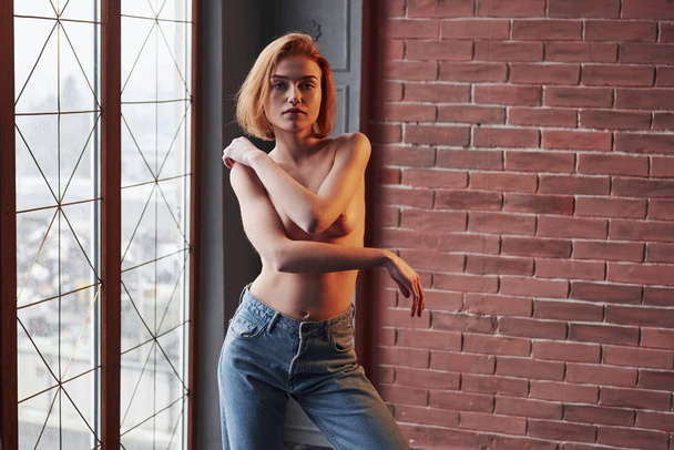 Uncensored photo. Hot young blonde with bare chest and jeans stands against the window and brick wall. - Φωτογραφία, εικόνα