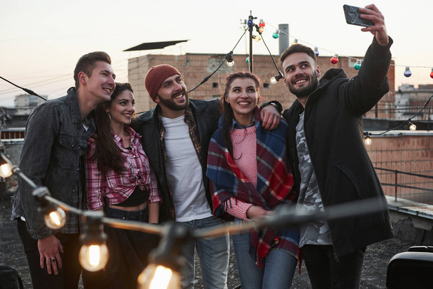 Look in the phone. Group of young cheerful friends having fun, hug each other and takes selfie on the roof with decorate light bulbs. - Φωτογραφία, εικόνα