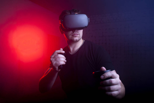 serious brutal guy with VR glasses stands in the virtual reality room and plays with joysticks, male gamer, virtual world - Photo, Image
