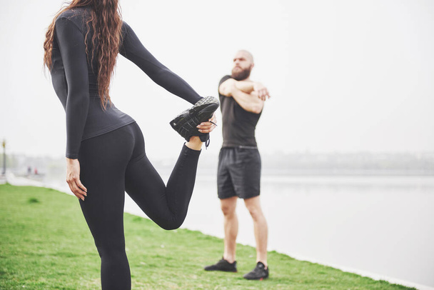 Fitness couple stretching outdoors in park near the water. Young bearded man and woman exercising together in morning. - Photo, Image