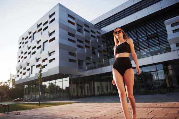 Sexy young woman in sunglasses and black bathing suit is standing on the city buildings background during sunset. - Fotografie, Obrázek